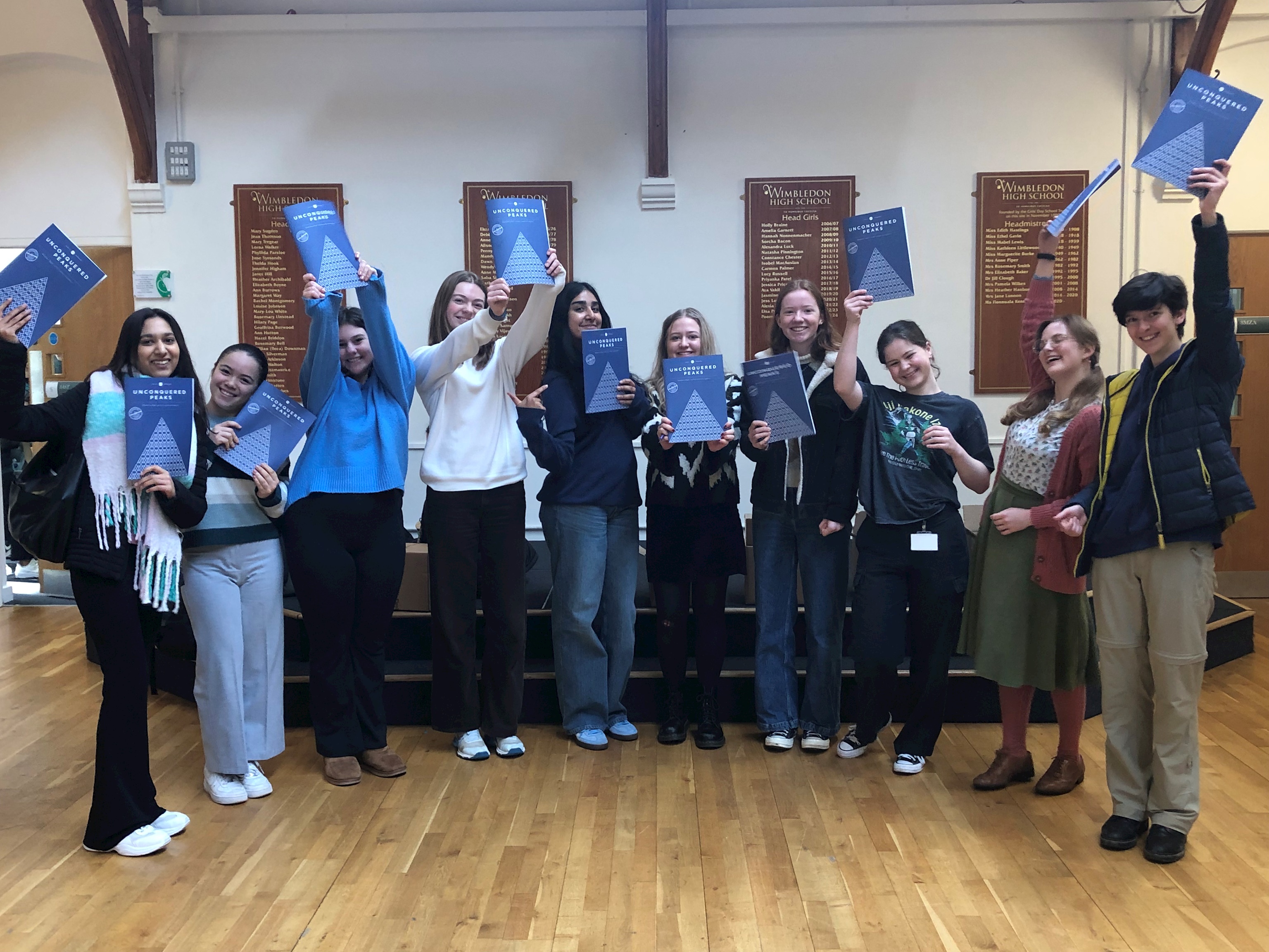 A photograph of all of the sixth form students who contributed to the latest edition of unconquered peaks. They are holding copies of the magazine in the air and looking happy