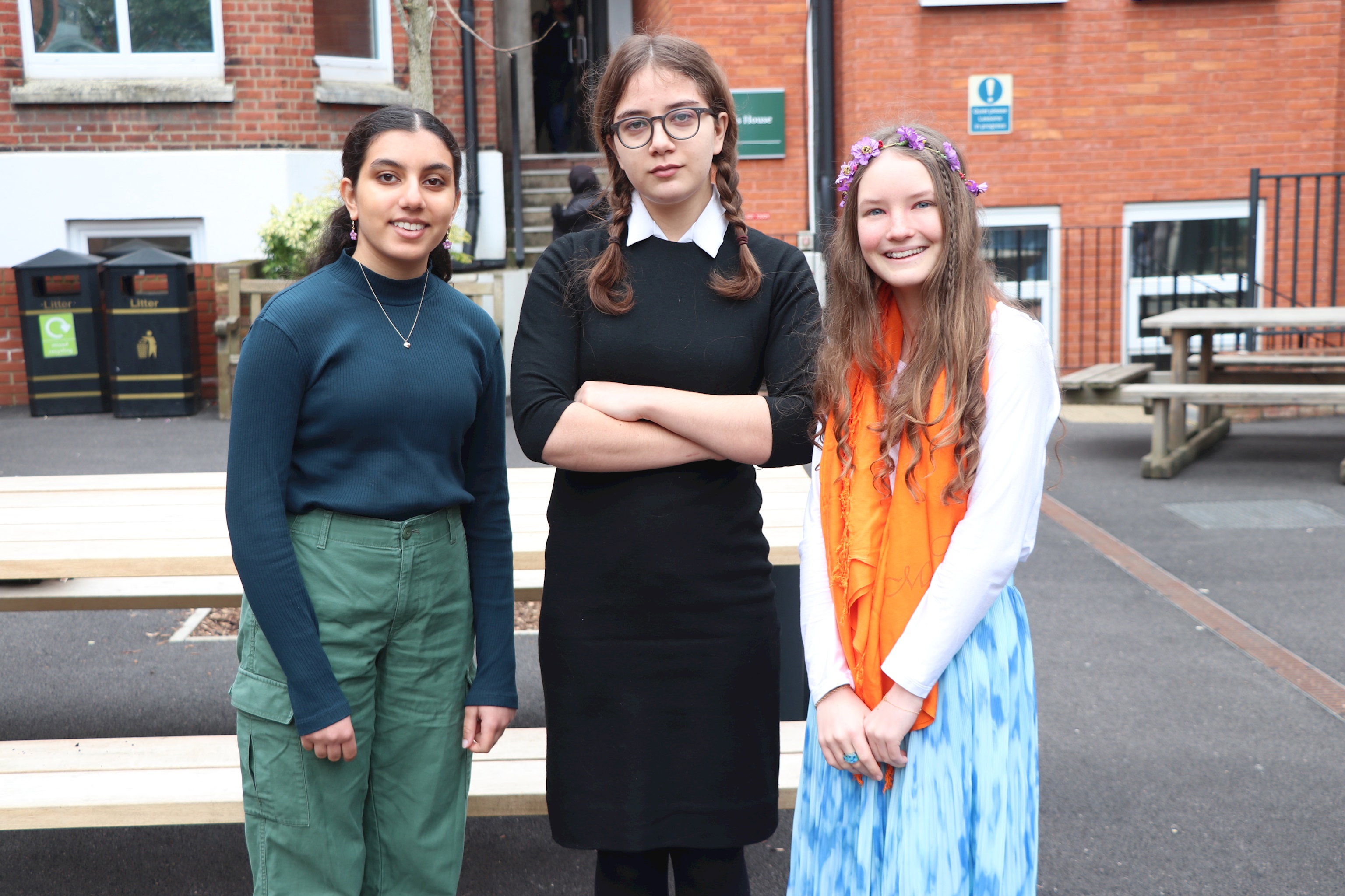 Three senior school girls dressed for World Book Day. The teenage girl on the left is dressed in green trousers a blue long-sleeved top; the girl in the middle is dressed in a black dress with braided hair; and the girl on the right is wearing a flowing blue skirt with an orange scarf and flowers in her hair