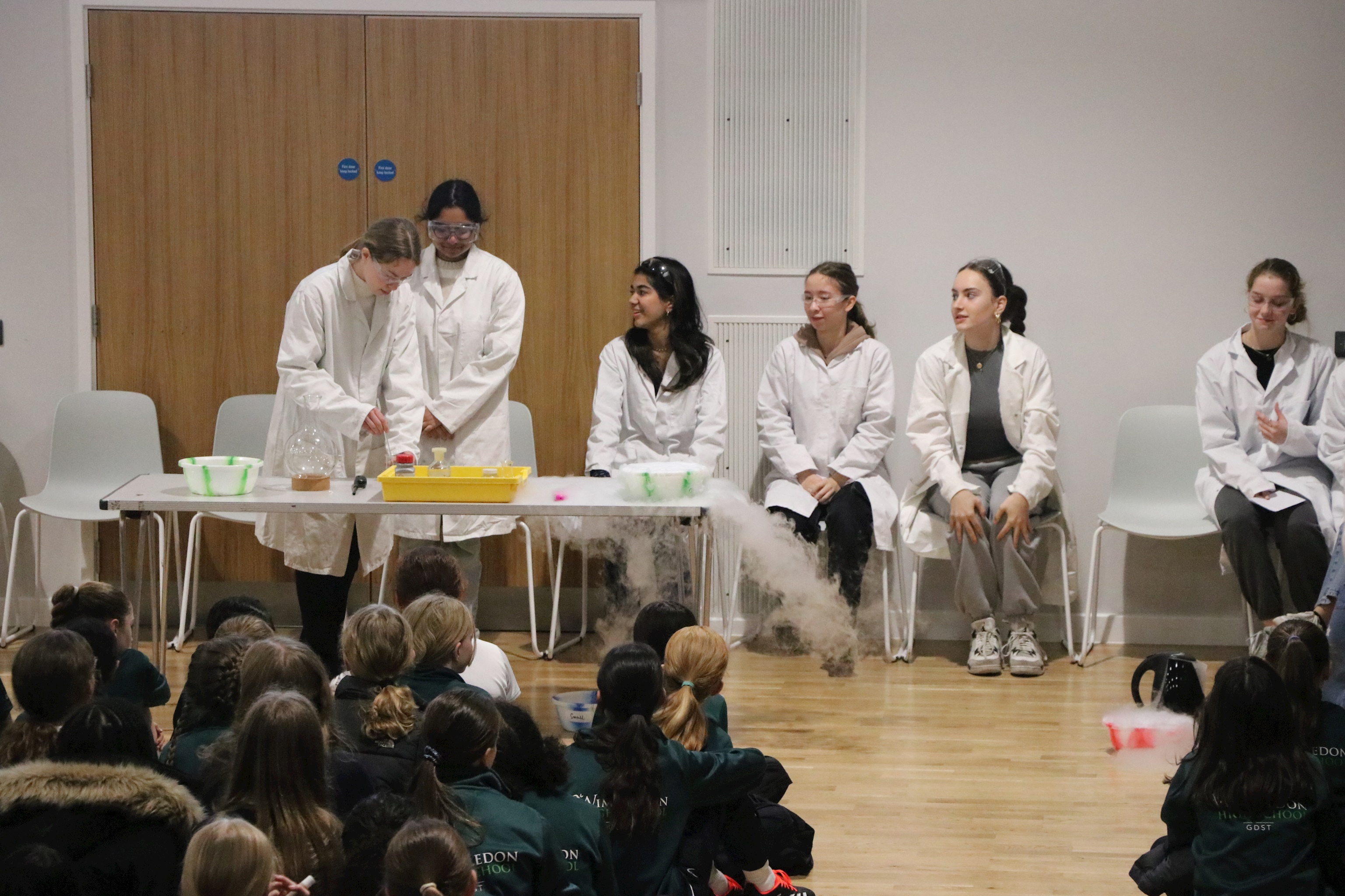 Six senior school girls wearing white lab coats and goggles. They are conducting an experiment with dry ice in front of the whole school during assembly