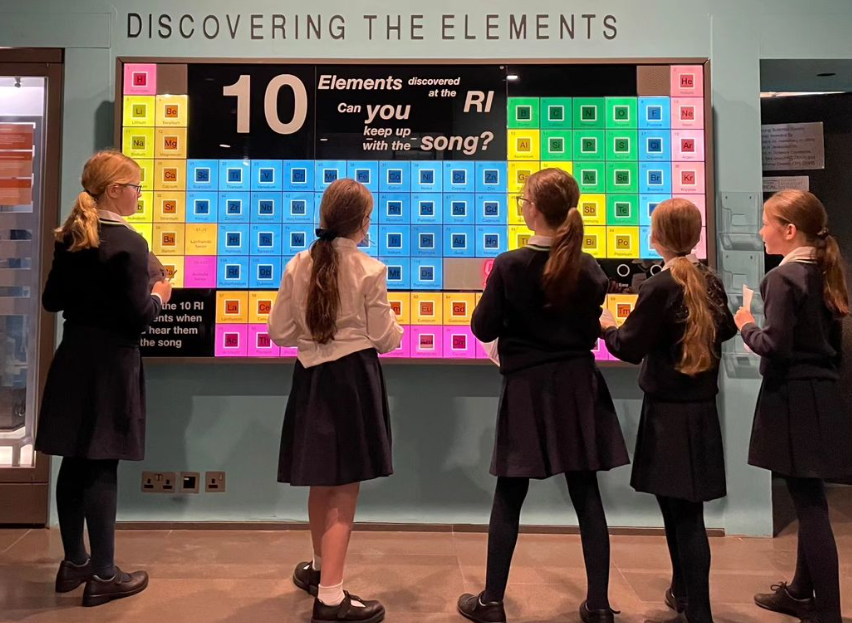Five Year 5 girls are standing in front of a large, colourful periodic table of elements that is projected on to a wall